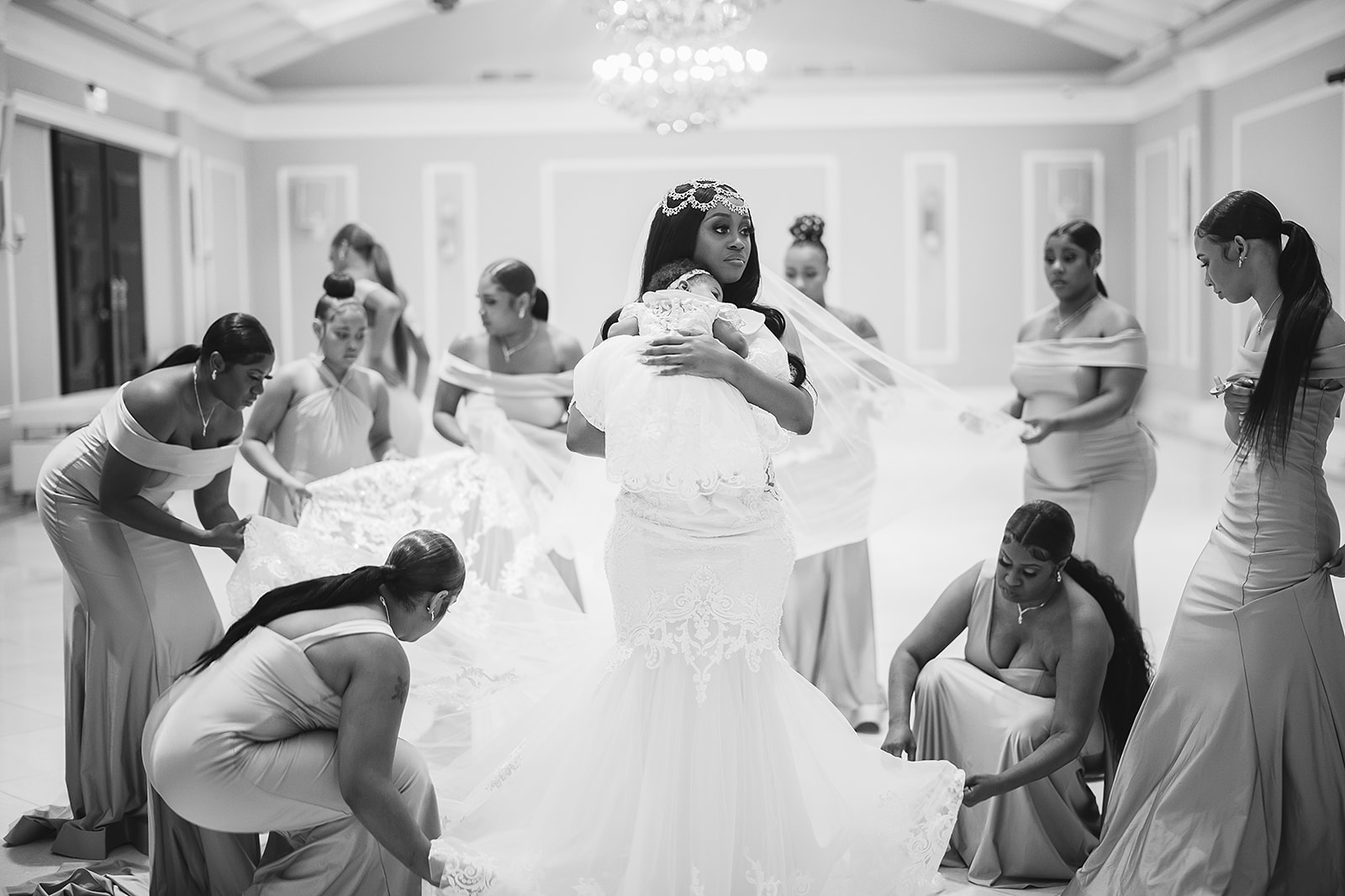 Bride holding baby while bridesmaids prepare dress in black and white