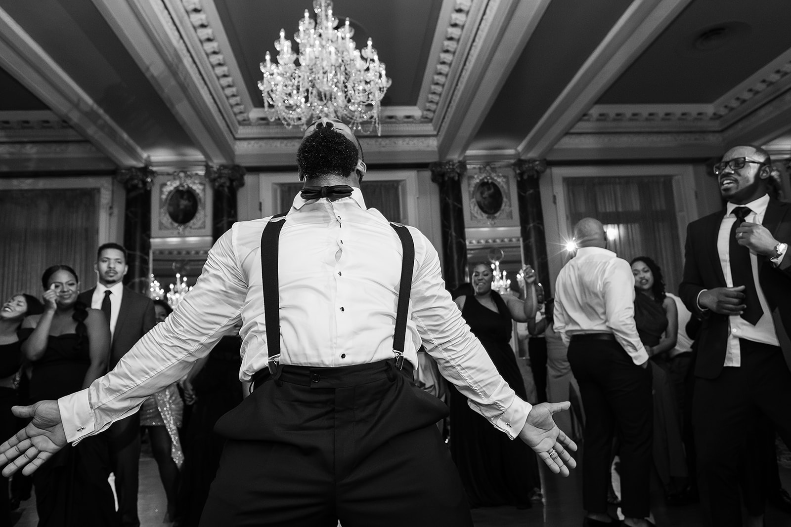 Groom dancing in suspenders on ballroom floor chandelier black and white