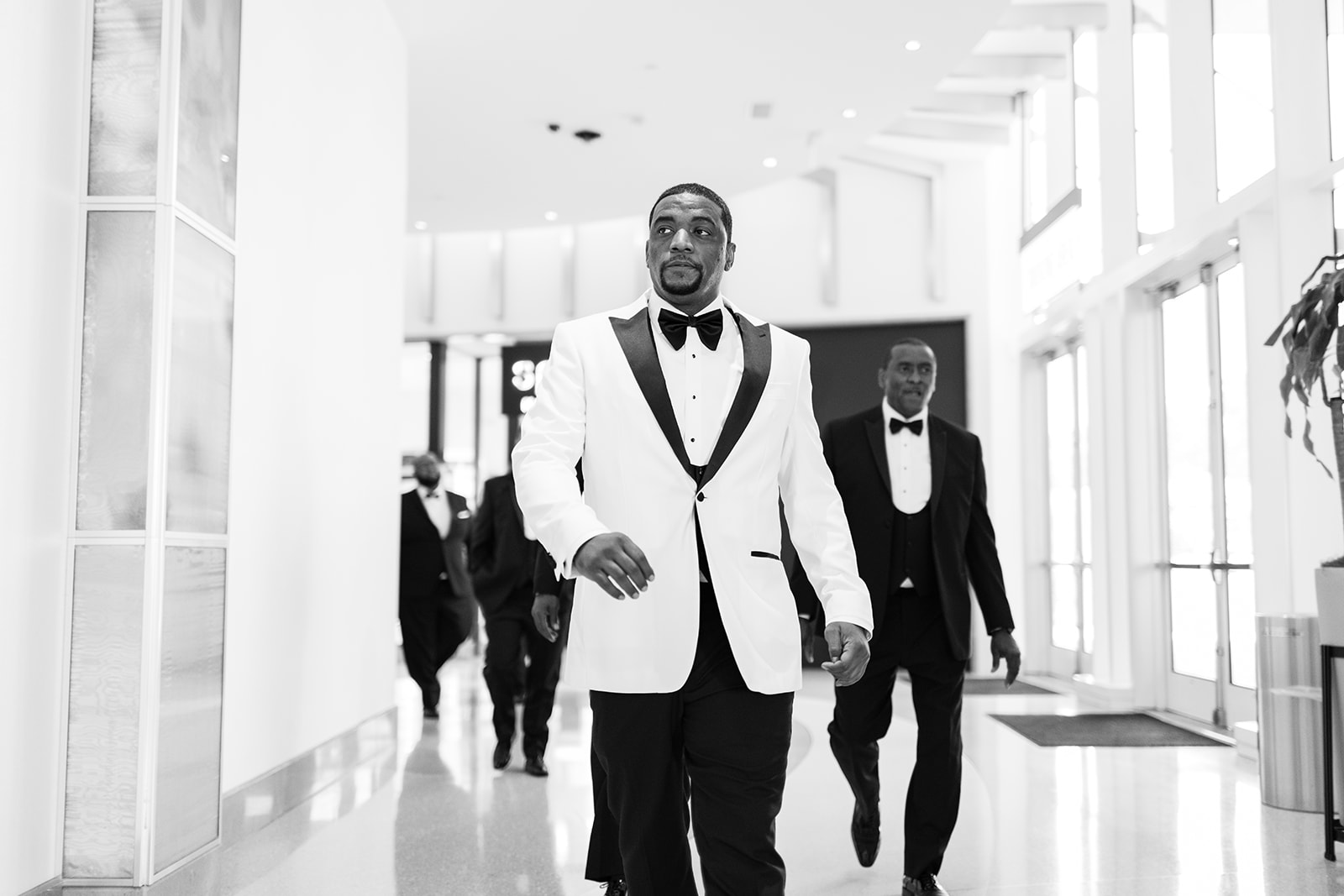 Groom in white jacket walking hallway with groomsmen black and white
